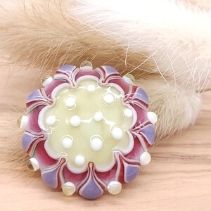 Könnte beinhalten: Eine lila, rote und weiße Glasperle mit einem Blumendesign und weißen Punkten. Die Perle ist handgefertigt und hat eine glänzende Oberfläche.