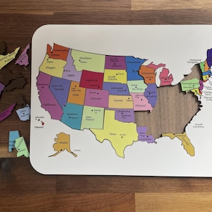 May include: A white board featuring a colorful map of the United States with individual state puzzle pieces. The map displays state names, capitals, and borders. Several detached state pieces are positioned to the left and right of the board.
