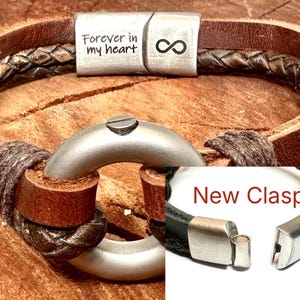 a leather bracelet with a metal clasp and a silver ring, both of which are attached to the bracelet. The bracelet is made of brown leather and has a braided pattern. The clasp is silver and has a metal ring attached to it. The bracelet is displayed on a wooden surface.