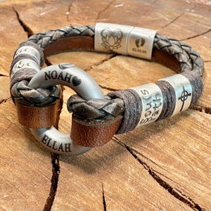 Puede incluir: Pulsera de cuero trenzado con detalles plateados. La pulsera presenta los nombres "Noah" y "Ellah" grabados en un cierre plateado. Cuentas plateadas adicionales est&aacute;n grabadas con texto y s&iacute;mbolos. La pulsera tiene una combinaci&oacute;n de colores marr&oacute;n y gris.