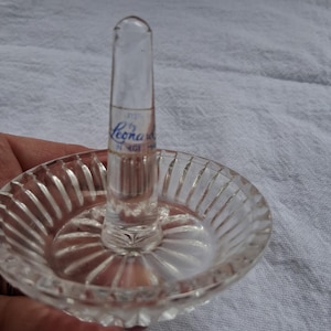 May include: A clear glass ring holder featuring a circular base with vertical fluted detailing and a central tapered post. The post displays a blue printed label that reads Leonard, with additional text below the name.