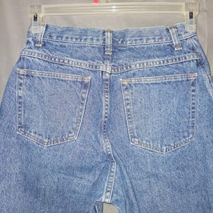 Może przedstawiać: Zbliżenie na tył niebieskich dżins&oacute;w. Dżinsy mają dwie tylne kieszenie i wiszą na przezroczystym plastikowym wieszaku. Denim ma średnie pranie z widocznymi szwami.