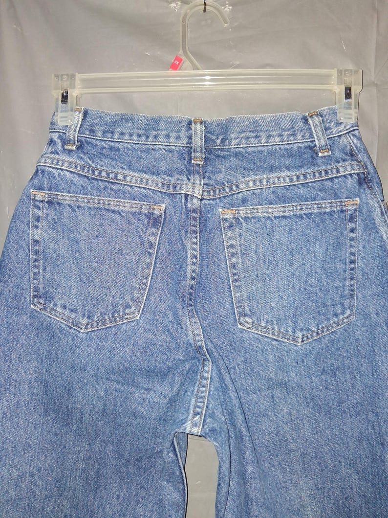 Może przedstawiać: Zbliżenie na tył niebieskich dżins&oacute;w. Dżinsy mają dwie tylne kieszenie i wiszą na przezroczystym plastikowym wieszaku. Denim ma średnie pranie z widocznymi szwami.