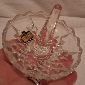 May include: A clear crystal ring holder dish featuring a central conical post for rings and a scalloped rim. The base has a faceted diamond-cut pattern. A small rectangular sticker on the surface reads CRYSTAL, 24% PbO, and Made in Germany.