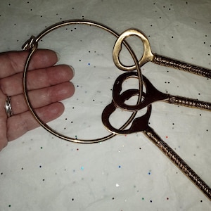 May include: A gold-colored metal bracelet with three decorative keys. The bracelet is a circular band with a clasp. The keys have ornate handles and heart-shaped keyholes. The background is white with colorful glitter.