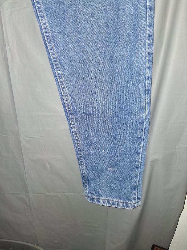 Może przedstawiać: Jasnoniebieskie dżinsy. Dżinsy mają klasyczny pięciokieszeniowy design z prostą nogawką. Denim ma średnie pranie z widocznymi szwami wzdłuż szw&oacute;w. Dżinsy są w stylu casual.