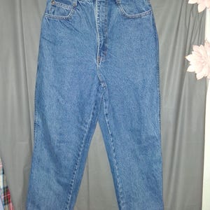 May include: Light blue denim jeans with a high waist, classic five-pocket design, and a button and zipper closure. The jeans are hanging on a white hanger against a gray backdrop. The jeans are a medium wash.