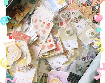 May include: A collection of colorful stickers in various shapes and sizes, including food, animals, and decorative elements. The stickers are packaged in clear plastic and arranged in a container. Some stickers have text, and the overall aesthetic is cute and playful.