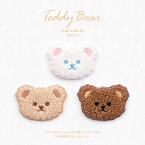 May include: Three embroidered teddy bear patches in white, beige, and brown. The white bear has pink ears and a blue nose and mouth. The beige bear has brown features, and the brown bear has beige ears and a brown nose and mouth. Text reads "Teddy Bear EMBROIDERY PATCH".