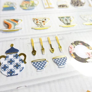 May include: A sheet of decorative stickers featuring various teacups, teapots, and utensils. The stickers showcase a variety of designs, including floral patterns, geometric shapes, and gold accents. The color palette includes blue, white, gold, and other pastel shades.