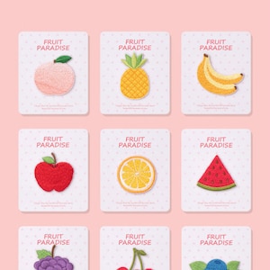 May include: Nine embroidered fruit patches on white cards with the text "FRUIT PARADISE". Fruits include a peach, pineapple, bananas, apple, orange slice, watermelon, grapes, cherries, and blueberries. The background is light pink.