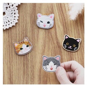May include: A collection of embroidered cat face patches in a range of colours, including white, orange, black, and grey. Each patch depicts a different cat expression, with details such as winking eyes and smiling faces. These patches are perfect for adding a playful touch to garments or accessories.