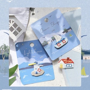 May include: Embroidered patches on blue cards with a nautical theme. One patch depicts a lighthouse, another a sailboat, and a third a house. The cards have the words "Lighthouse" and "Ocean Ship" printed on them, with a background of a seascape.