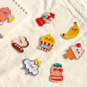 May include: A selection of colourful embroidered patches with food and whimsical character designs. Includes a gumball machine, banana, ice cream cone, popcorn, bread, cloud with star, and a drink with the word "Yummy".