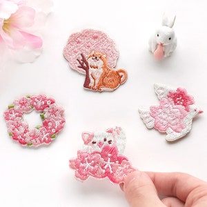 May include: Embroidered patches in pink, white, and brown. Designs include a Shiba Inu under a pink tree, a floral wreath, a white bunny, a bird with pink flowers, and a bunny with cherry blossoms. Ideal for clothing or craft projects.
