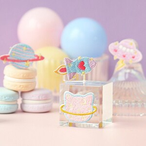 May include: A collection of embroidered space-themed patches in pastel colours. The patches include a rocket, a planet, a cat, and a cloud with lightning. The patches are displayed on a clear cube, with macarons in the background.