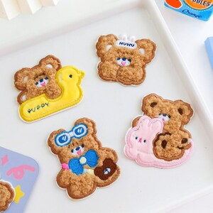 May include: Embroidered patches with teddy bear designs. One bear is on a yellow duck, another wears glasses and a bow tie, and a third has a bunny. The patches are brown, white, blue, and pink, perfect for clothing or accessories.
