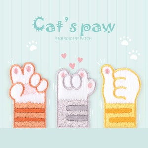 May include: Three embroidered cat paw patches in orange, grey, and yellow, each with white paws and pink accents. The patches are in the shape of hand gestures: peace sign, thumbs up, and a fist. The text "Cat's paw" and "Embroidery patch" are at the top.