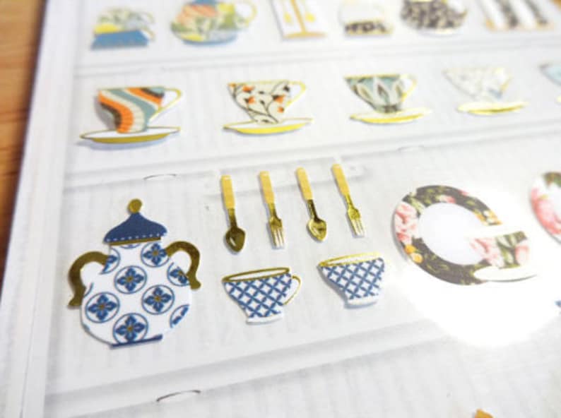 May include: A sheet of decorative stickers featuring various teacups, teapots, and utensils. The stickers showcase a variety of designs, including floral patterns, geometric shapes, and gold accents. The color palette includes blue, white, gold, and other pastel shades.