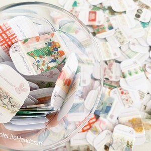 May include: A clear glass jar filled with numerous small, decorative paper samples. The samples feature various patterns, including floral designs, bunnies, and grid patterns. The text "Mini Sample" and "ASimpleLifeHandcraft" are visible on some samples.