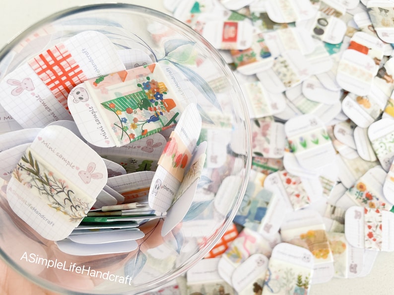 May include: A clear glass jar filled with numerous small, decorative paper samples. The samples feature various patterns, including floral designs, bunnies, and grid patterns. The text "Mini Sample" and "ASimpleLifeHandcraft" are visible on some samples.