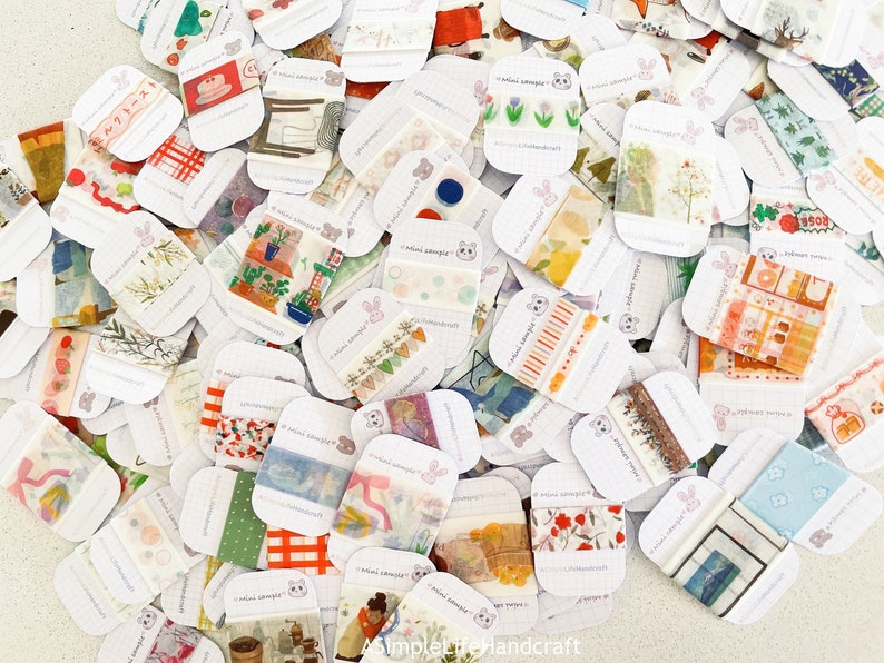 May include: A collection of decorative washi tape samples, each mounted on a small white card. The tapes feature various designs, including floral patterns, food illustrations, and geometric shapes. The cards are scattered across a light-colored surface.