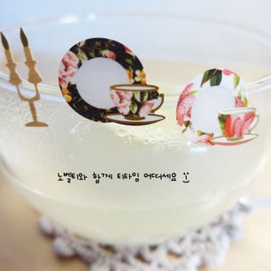 May include: A clear glass mug filled with a light-colored liquid, possibly tea or lemonade. Decorative stickers of teacups and saucers with floral patterns are placed on the mug. The text in the image reads "노벨티와 함께 티타임 어떠세요 :) ".