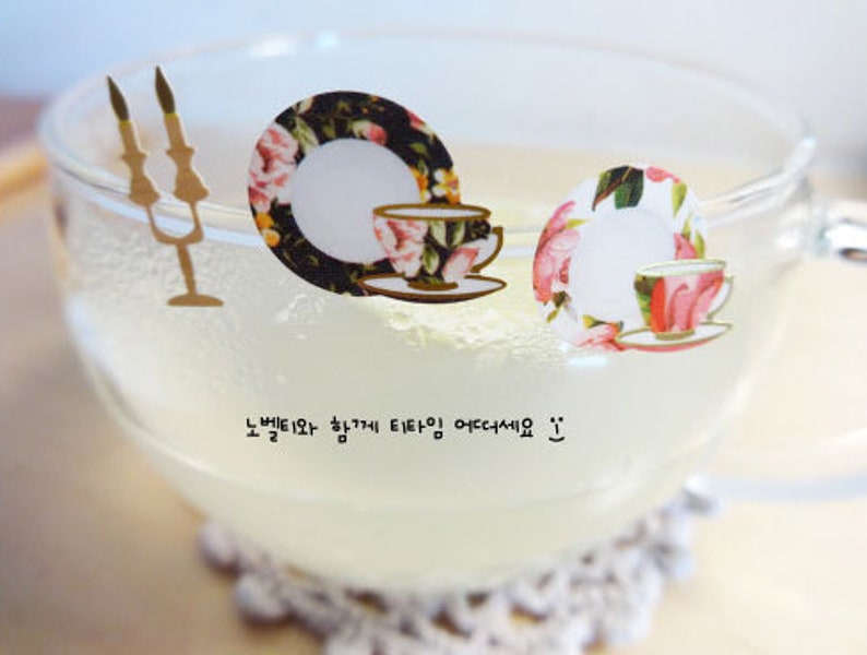 May include: A clear glass mug filled with a light-colored liquid, possibly tea or lemonade. Decorative stickers of teacups and saucers with floral patterns are placed on the mug. The text in the image reads "노벨티와 함께 티타임 어떠세요 :) ".