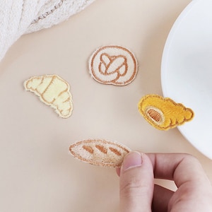 May include: Embroidered patches of baked goods, including a croissant, pretzel, and loaf of bread. The patches are in shades of beige, tan, and yellow, with detailed stitching. A hand is holding one of the patches.