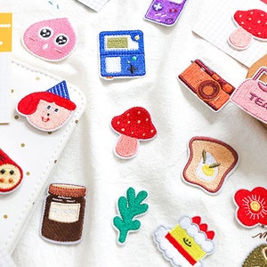 May include: A selection of embroidered patches with diverse designs. Includes a red mushroom, a retro video game, a slice of toast with an egg, a cake, a camera, and a jar of jam. Some patches have text.