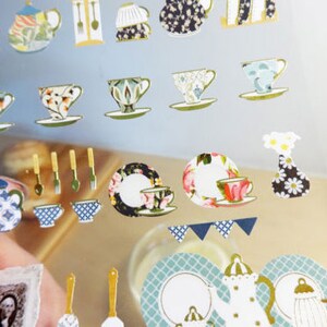 May include: A sheet of decorative stickers featuring various teacups, teapots, and serving utensils. The stickers have a vintage aesthetic with floral and patterned designs in colors like blue, green, and gold. Some stickers include small plates and a vase.