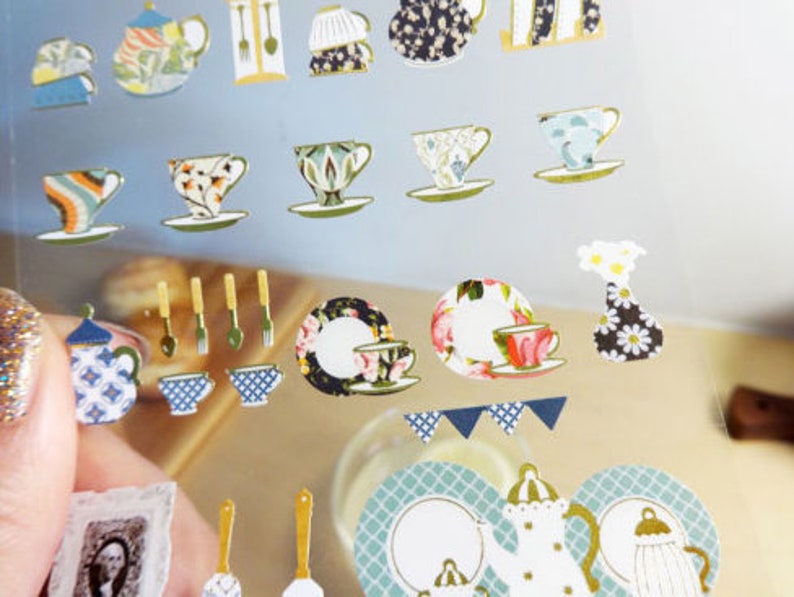 May include: A sheet of decorative stickers featuring various teacups, teapots, and serving utensils. The stickers have a vintage aesthetic with floral and patterned designs in colors like blue, green, and gold. Some stickers include small plates and a vase.