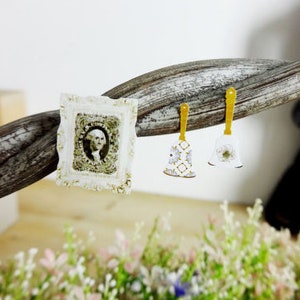 May include: A small, ornate white frame with a portrait of a person, and two bell-shaped earrings with gold handles. One earring has a floral pattern, the other a flower. The items are displayed on a twisted wooden branch, with greenery in the foreground.