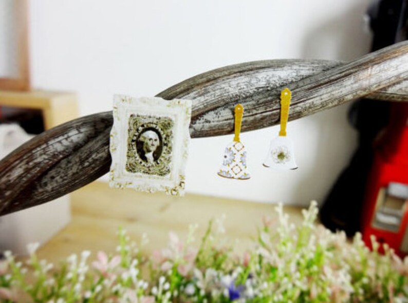 May include: A small, ornate white frame with a portrait of a person, and two bell-shaped earrings with gold handles. One earring has a floral pattern, the other a flower. The items are displayed on a twisted wooden branch, with greenery in the foreground.
