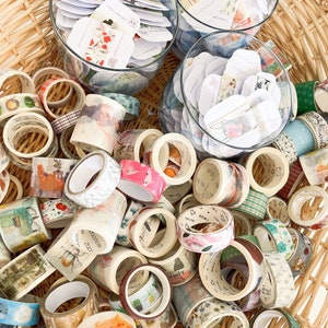May include: A wicker basket overflowing with decorative washi tape rolls in various colors and patterns. Clear glass bowls hold small, decorative tags. The tape designs include floral, geometric, and illustrated themes, creating a colorful and artistic display.