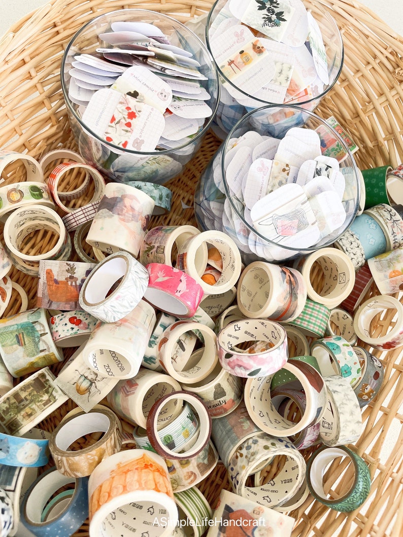 May include: A wicker basket overflowing with decorative washi tape rolls in various colors and patterns. Clear glass bowls hold small, decorative tags. The tape designs include floral, geometric, and illustrated themes, creating a colorful and artistic display.