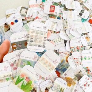 May include: A collection of mini sample washi tapes with various patterns, including floral, geometric, and illustrated designs. Each tape is displayed on a white card with a grid pattern and the text "Mini sample". The tapes are held in a hand.