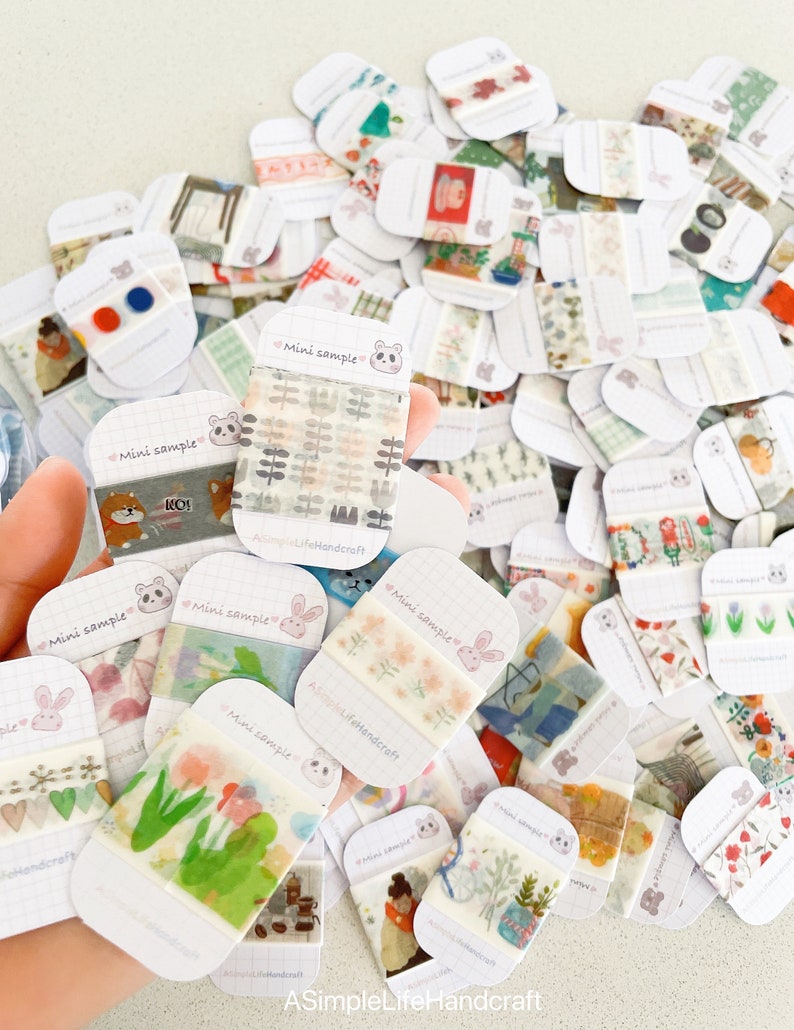 May include: A collection of mini sample washi tapes with various patterns, including floral, geometric, and illustrated designs. Each tape is displayed on a white card with a grid pattern and the text "Mini sample". The tapes are held in a hand.