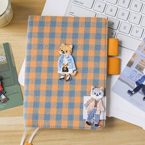 May include: A notebook with an orange and blue checked cover, decorated with embroidered cat figures. The notebook is open on a laptop keyboard, with stickers and pencils nearby. The text "喵咪穿搭手册" is visible.