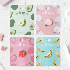 May include: Four fruit-themed pins on decorative cards. The pins feature an avocado, peach, strawberry, and banana. Each card has a matching fruit pattern and Chinese text. The pins are small and detailed.