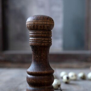 May include: A vintage wooden pepper mill with a dark brown finish. The pepper grinder has a rounded top and a bulbous body, with a base. The mill is standing on a weathered wooden surface.