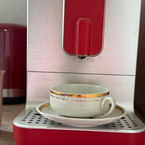 May include: A silver and red coffee machine with a red vertical panel and a drip tray. A white ceramic teacup with a gold rim and matching saucer sits on the drip tray. The cup has a curved handle and a smooth finish, contrasting with the metallic machine.