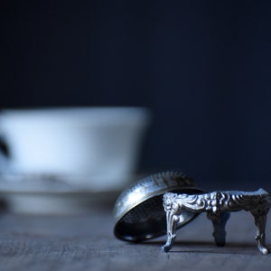 Puede incluir: Un dedal de plata y una pequeña mesa de plata son el foco de esta imagen. El dedal está volcado, revelando su intrincado diseño. Una taza y un platillo blancos están borrosos en el fondo, lo que sugiere un ambiente temático de té.
