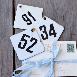 May include: Three white, square metal tags with black numbers 91, 34, and 52. A bundle of vintage postcards tied with a light blue lace ribbon is below the tags. The background is a weathered wooden surface.