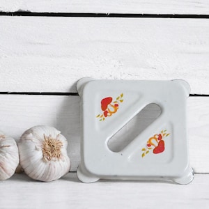 May include: A square white metal trivet with rounded corners and a diagonal slot in the center. The surface features a small graphic of red mushrooms with green leaves on two corners. Two whole garlic bulbs are positioned to the left of the trivet.