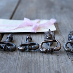 Puede incluir: Cuatro tiradores de cajón antiguos en tono bronce con diseños ornamentados, dispuestos sobre una superficie de madera envejecida. Una pequeña tarjeta con una cinta y encaje rosa añade un toque de elegancia. Ideales para restaurar muebles antiguos.