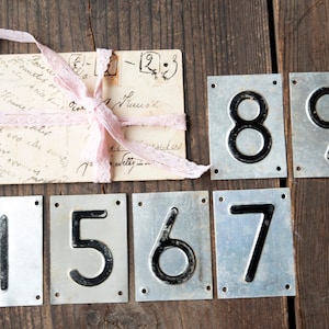 Könnte beinhalten: Vintage Metallnummernschilder mit schwarzen Ziffern, angeordnet auf einer Holzoberfläche. Die Zahlen 1, 5, 6, 7, 8 und 9 sind abgebildet. Eine antike Postkarte, die mit einem rosa Spitzenband gebunden ist, ist ebenfalls vorhanden.