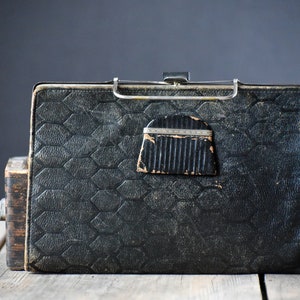 Op de afbeelding: Een antieke zwarte leren koffer met een geometrisch patroon en metalen sluiting. De koffer heeft een rechthoekige vorm en een kleine, decoratieve flap. De koffer vertoont tekenen van slijtage, wat suggereert dat het een oud item is.