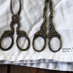 Puede incluir: Dos pinzas de servir de plata antiguas con diseños florales ornamentados, descansando sobre una servilleta de lino blanco con el texto "stone washed pure linen."