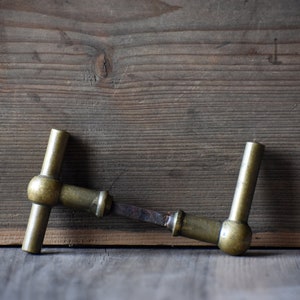 Könnte beinhalten: Antiker Türgriff aus Messing mit T-förmigem Design. Der Griff weist Alters- und Gebrauchsspuren auf, mit einem leicht verrosteten Verbindungsstück. Der Hintergrund ist eine rustikale Holzoberfläche.